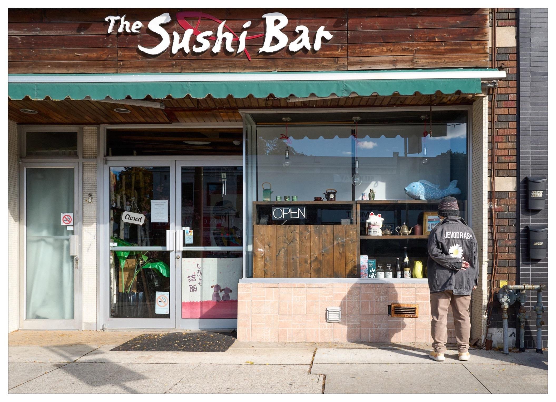 Storefront of "The Sushi Bar" with a wooden and dark brick exterior. The entrance features a glass door with a "Closed" sign, flanked by a large window displaying a blue fish sculpture and various items. A person in winter clothing stands reading a menu by the window, marked by an "Open" sign on the countertop.