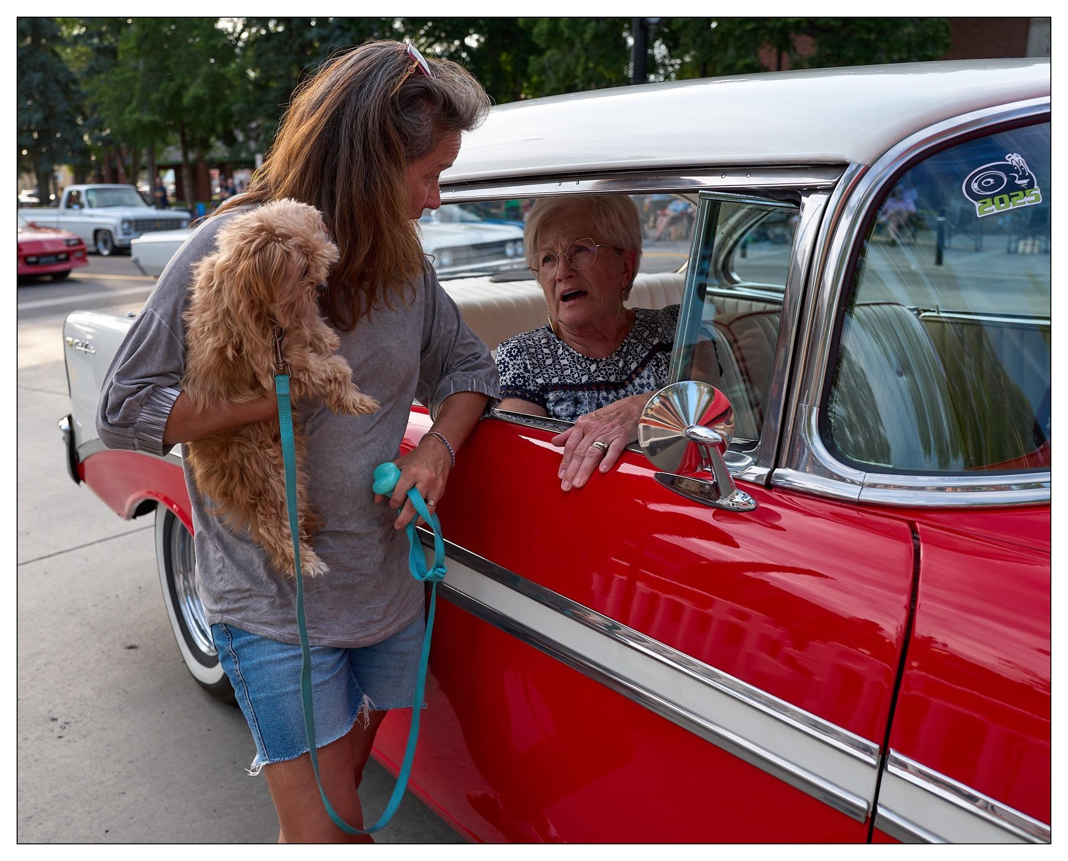 A woman holding a small brown dog engages with an elderly woman seated in a bright red vintage car. The car features chrome accents and a side mirror. They are on a street, with other cars visible in the background.