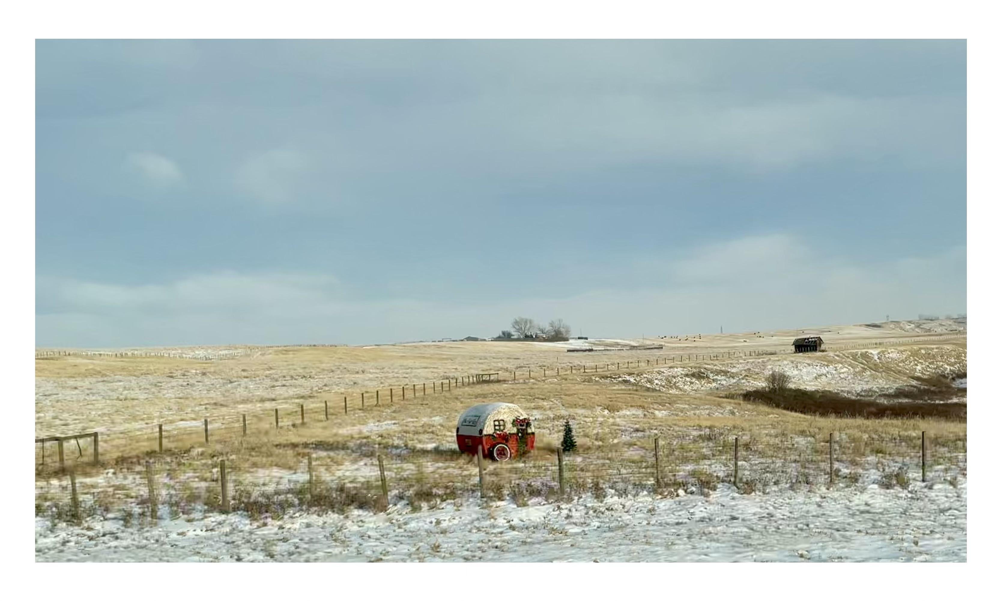 A rural landscape covered in patches of snow with a clear sky above. A red vintage camper decorated with a wreath is parked in a fenced field. Actually, it is a round bale with a cover painted to look like a small round camper trailer. 