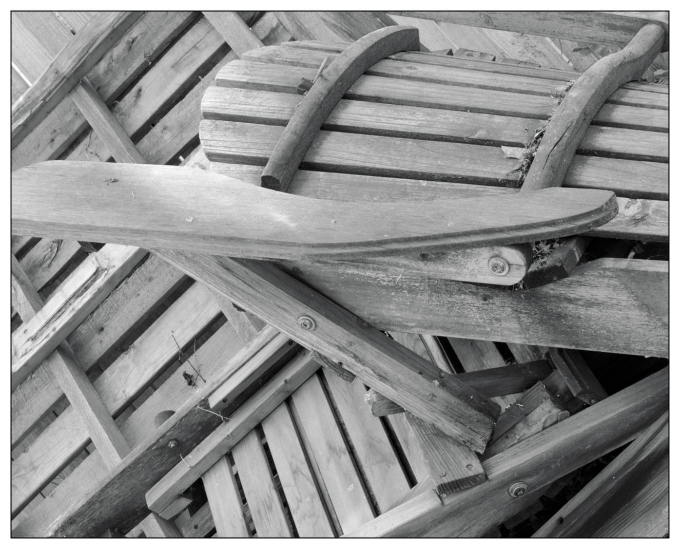 Black and white photograph of a pile of wooden chairs stacked haphazardly. The visible chair on top features curved back slats and armrests, with weathered and chipped wood textures prominently displayed. The underlying chairs are mostly obscured, showing only portions of their wooden slats.