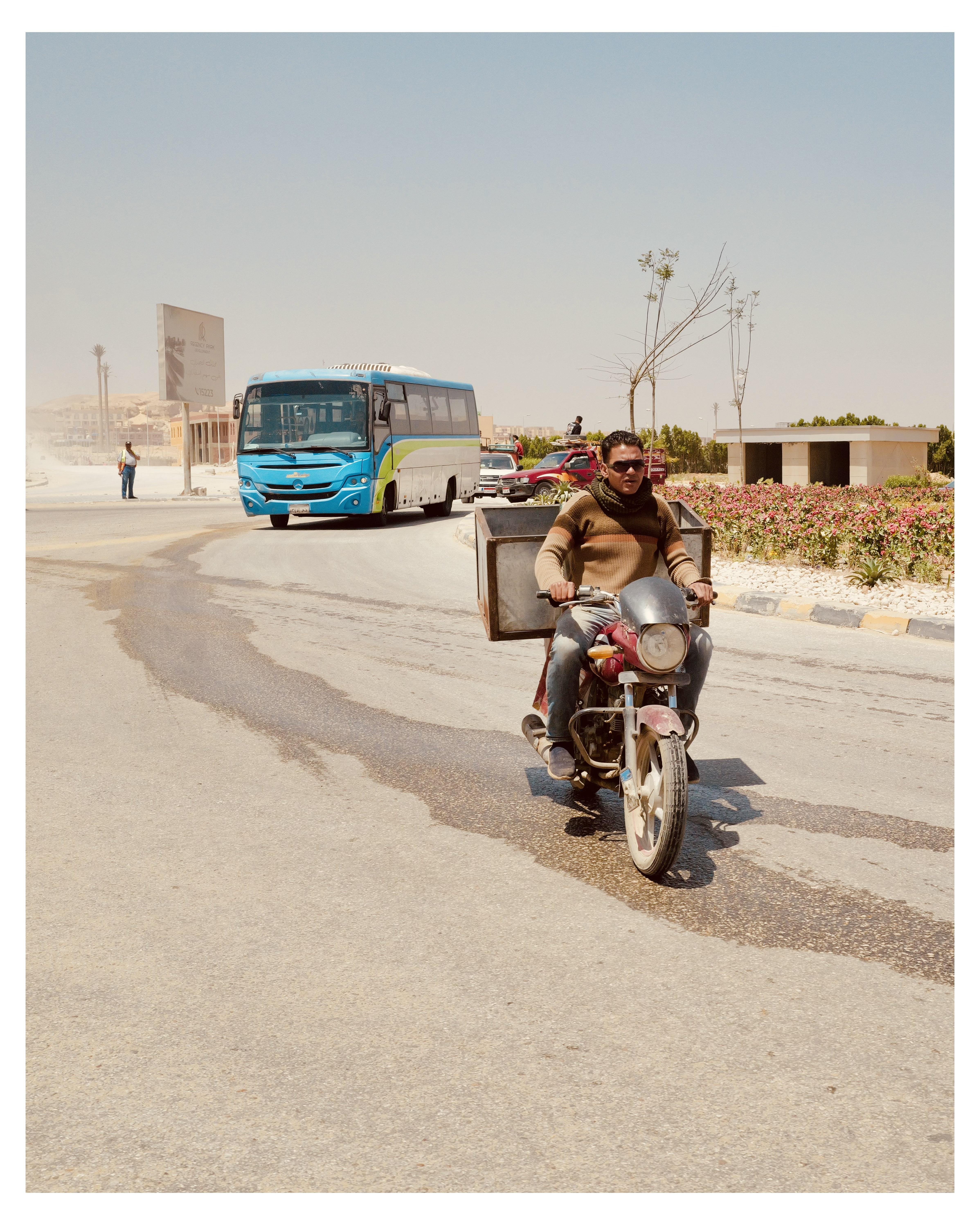 This image depicts a street scene, likely in a warm, arid place judging by the landscape and the brightness of the sunlight. In the foreground, there is a man riding a motorcycle, carrying a large item in a container attached to the back of his bike. He wears casual clothing and sunglasses. Behind him, there is a brightly colored blue and green bus on the road, indicating a form of public transportation. The surroundings include a relatively barren landscape with low bushes and a few trees, and some modern structures in the background. This setting suggests a mix of urban and semi-urban elements, likely on the outskirts of a city.