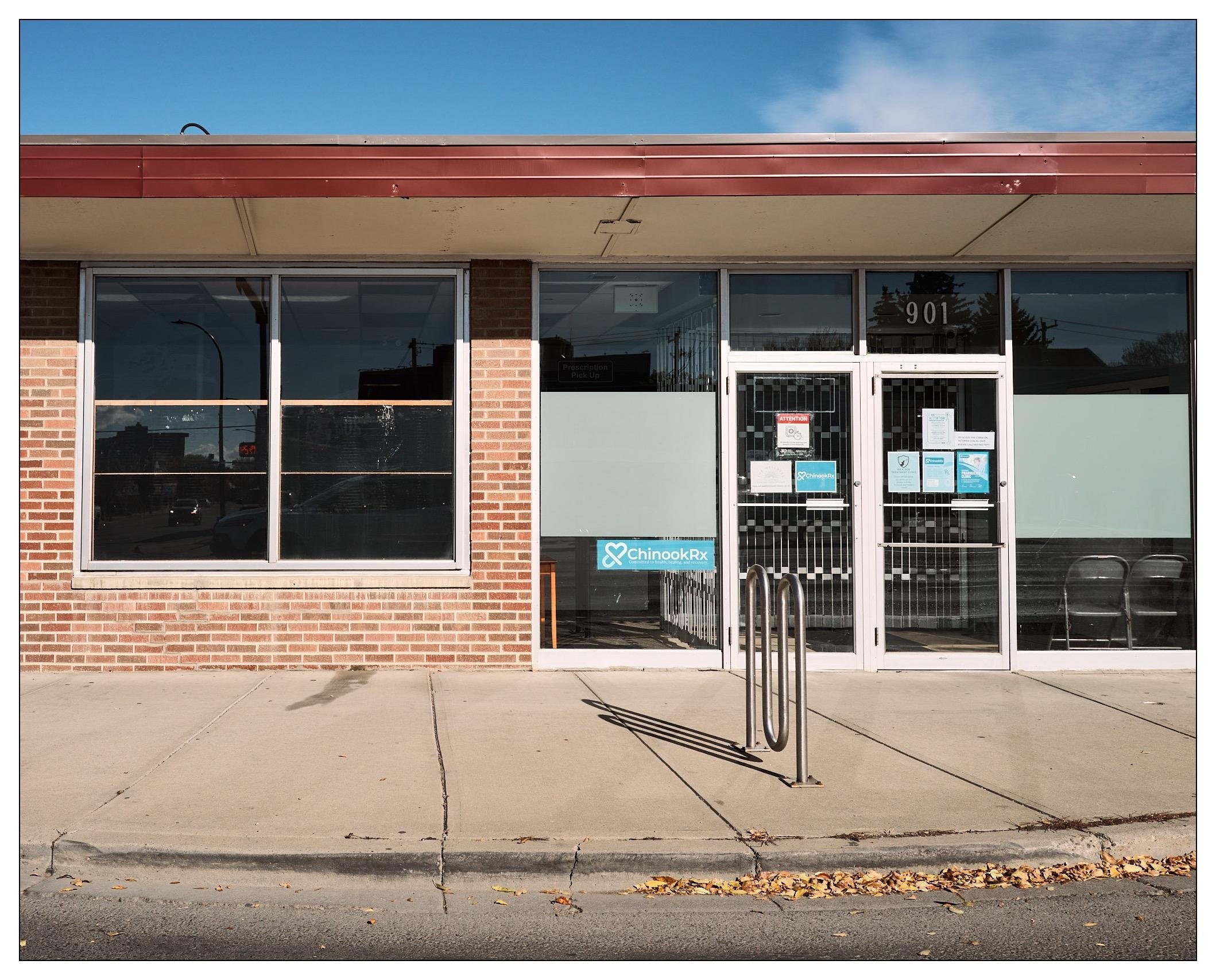 Exterior view of a single-story, brick facade building identified as "ChinookRx" with address number 901. It features a central glass door flanked by large windows and metallic door handles, with signage and notices attached to the door. In front, there is a metal bike rack and two folding chairs visible through the door's glass. The building has a red overhang and a clear blue sky is visible above.