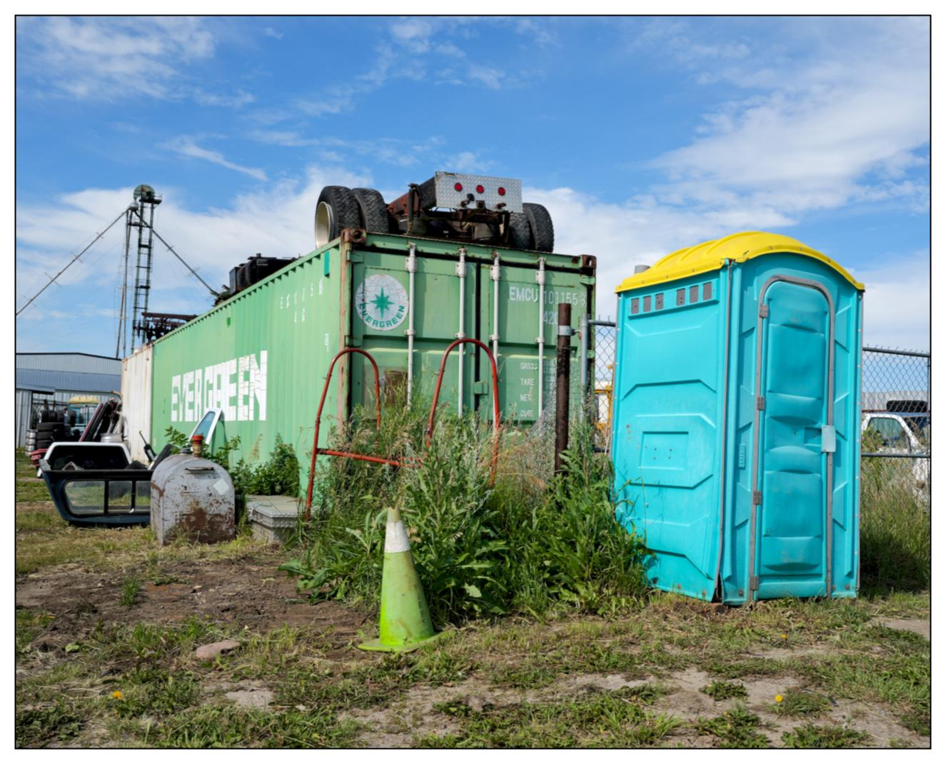 Green waste bin and blue portable toilet stand next to each other in a grassy area with a partial view of a vehicle and industrial structures in the background. The waste bin has rust and graffiti, and there are a car door and a traffic cone on the ground near it.
