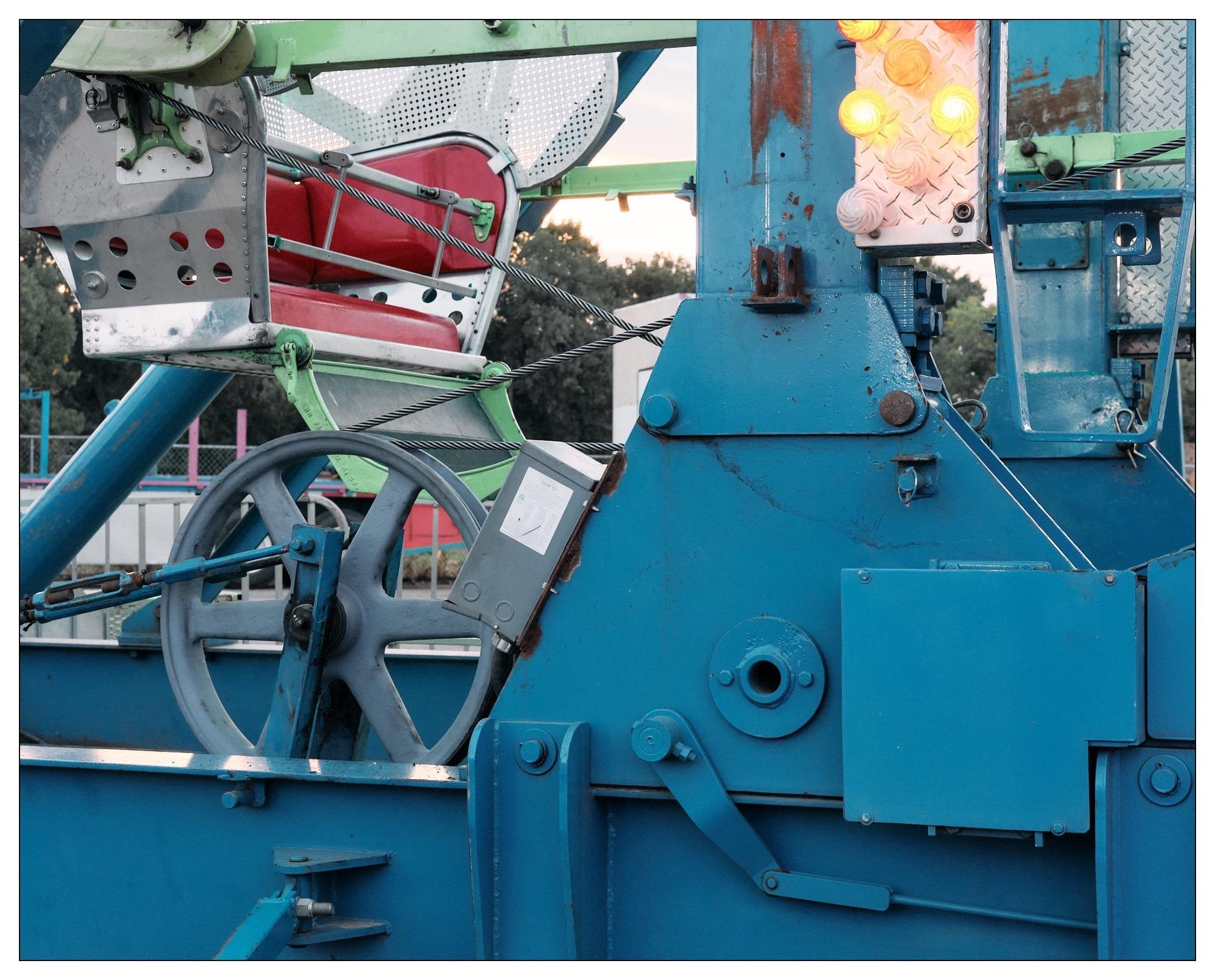 The image shows a close-up view of a part of a ferris wheel, specifically focusing on its mechanical components and machinery. There's a visible red seat with a security barrier, part of the ride's structure. The main body is painted in blue, with various mechanical elements like gears, levers, and steel framework shown in detail. There are also some illuminated lights visible, adding a festive or operational aspect to the machinery. The setting seems to be in the evening or dusk, given the lighting and sky in the background.