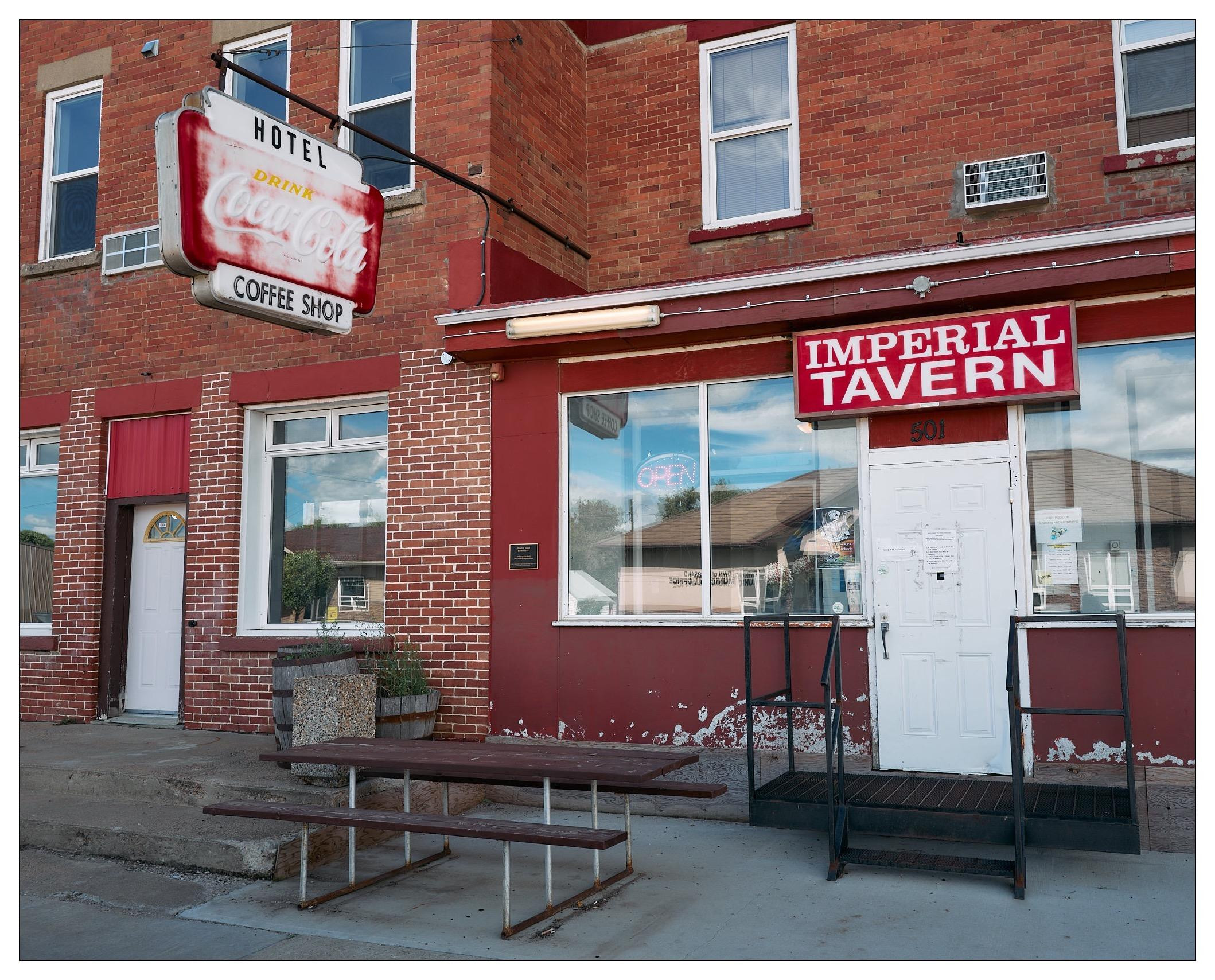 Red brick building corner with "Imperial Tavern" sign over white double doors marked 501. Adjacent is another entrance under a faded red "Hotel - Drink Coca-Cola - Coffee Shop" sign, next to a wooden bench on a sidewalk. Clear sky and a reflective window showing building reflections. Peeling white paint visible on the lower wall.