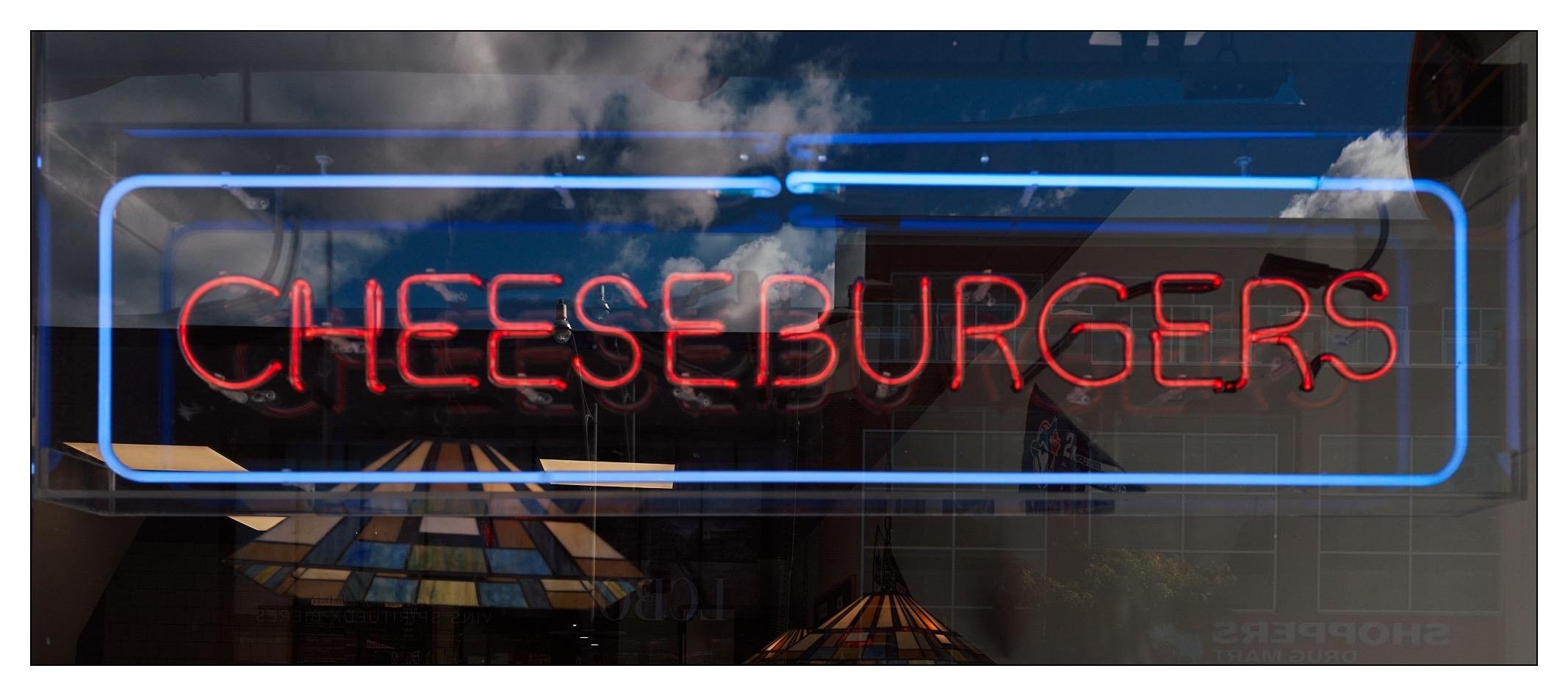 Neon sign reading "CHEESEBURGERS" in red, encased in a blue rectangular border, against a reflective window showing clouds and the interior ceiling of a restaurant.