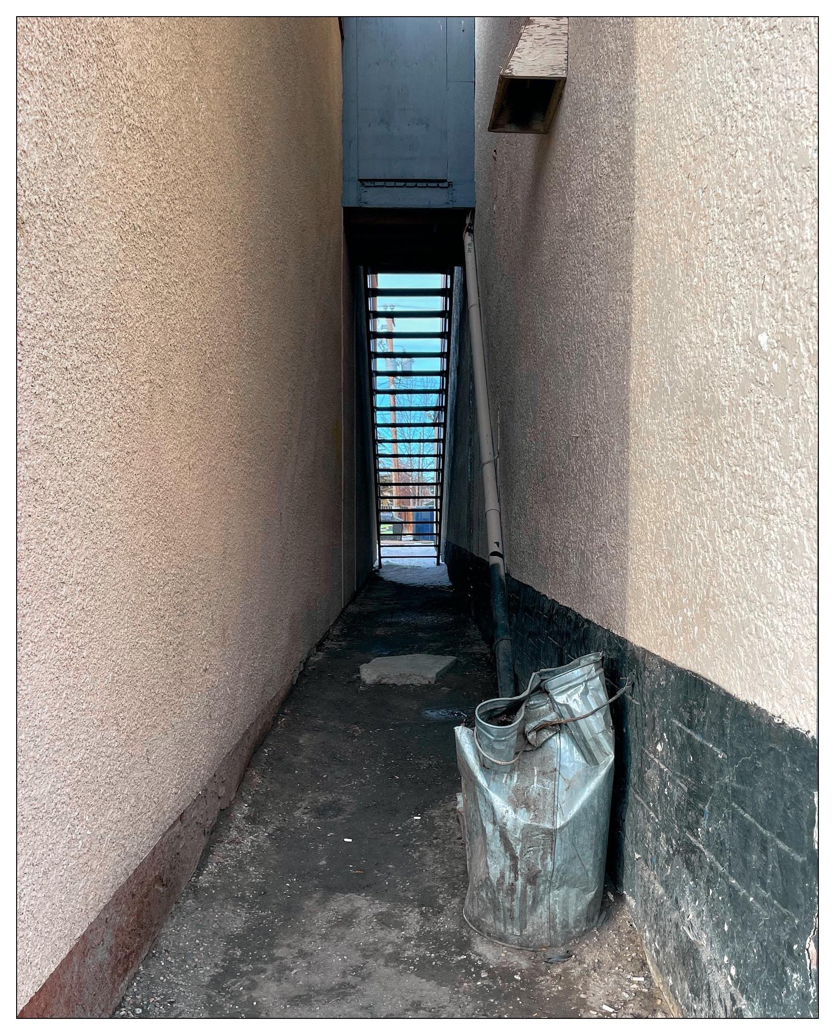 A very narrow alleyway between two buildings. The walls are textured with some visible stains or water marks. On the ground, there are some spots and debris indicating that the area might not be regularly cleaned. To the right, there is a traditional metal trash can, secured to the right hand wall with a cable.   The trash can is mangled, bent, twisted.   It is trashed.   At the far end of the alley, there's a set of open stairs letting in light from beyond this alley.