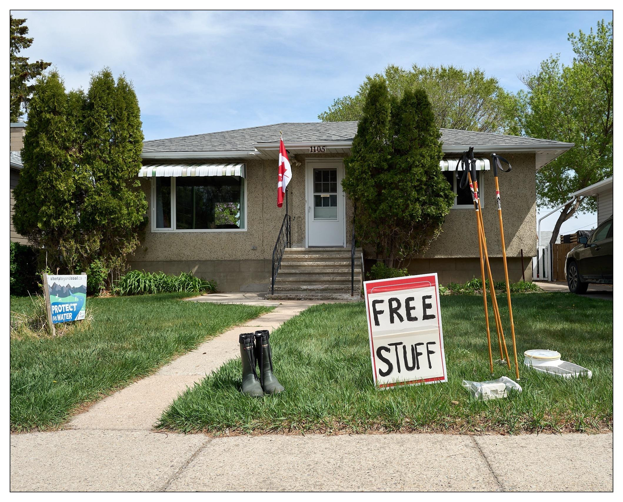 The image shows the front of a house, which appears to be a bungalow with a textured facade and a Canadian flag hanging by the door. On the lawn there are several items apparently offered for free, as indicated by a "FREE STUFF" sign. The items include a pair of black boots, a floor scale, and some walking poles standing vertically next to the sign. There is also a "PROTECT OUR WATER" yard sign suggesting environmental advocacy. The scene is set in a residential area with trees and a walkway leading to the house.