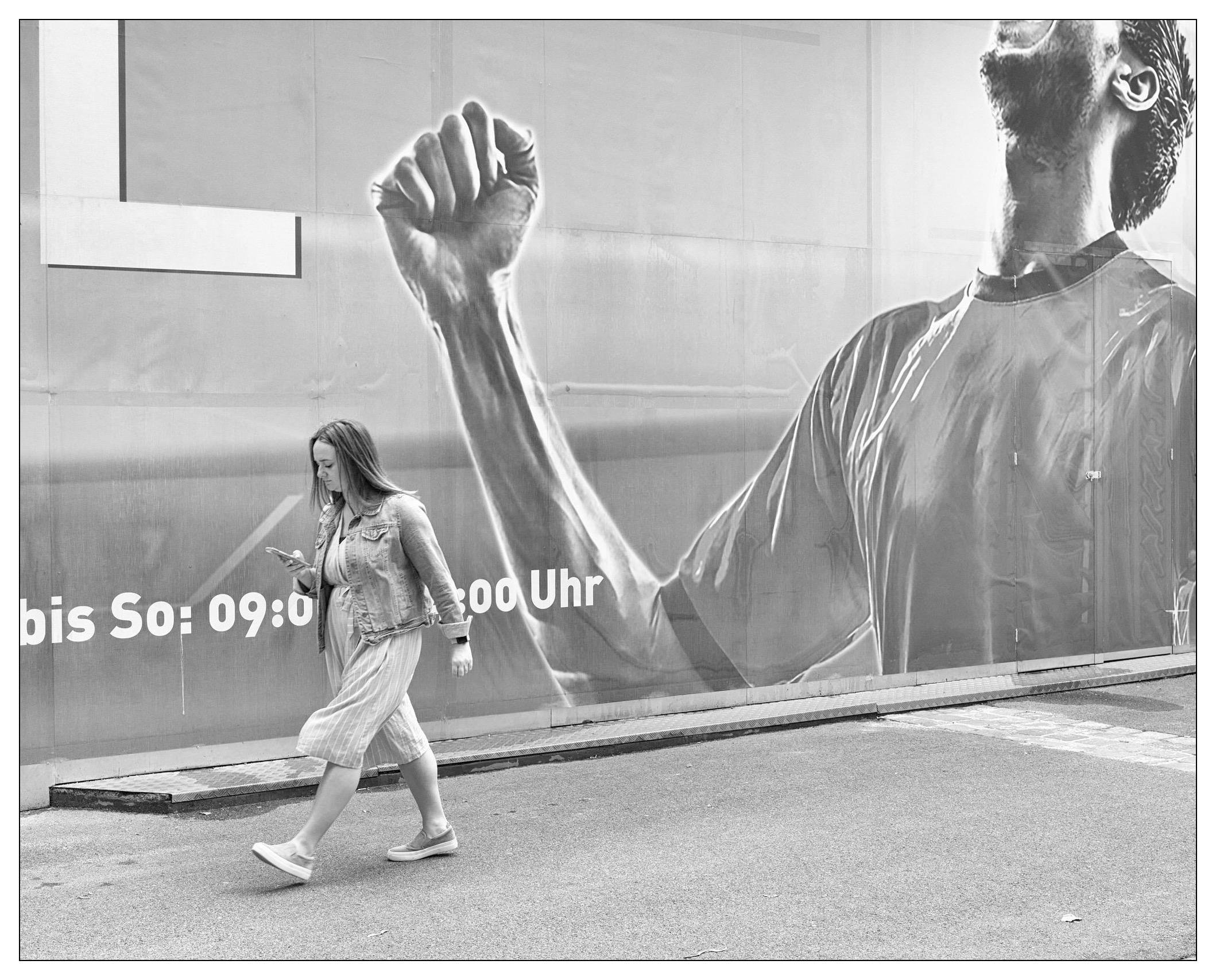 The image features a woman walking past a large outdoor advertisement. The advertisement depicts a man with a clenched fist raised high, suggesting a motif of strength or resistance. The billboard has a part with text in German, which reads "bis So: 09:00 - 20:00 Uhr," indicating business hours from Monday to Sunday, from 09:00 to 20:00 hours. This setting seems to be in an urban environment, given the large advertising panel and the street context implied by the woman's casual stroll and use of a smartphone. The entire photograph is in black and white, which adds a timeless quality to the scene.