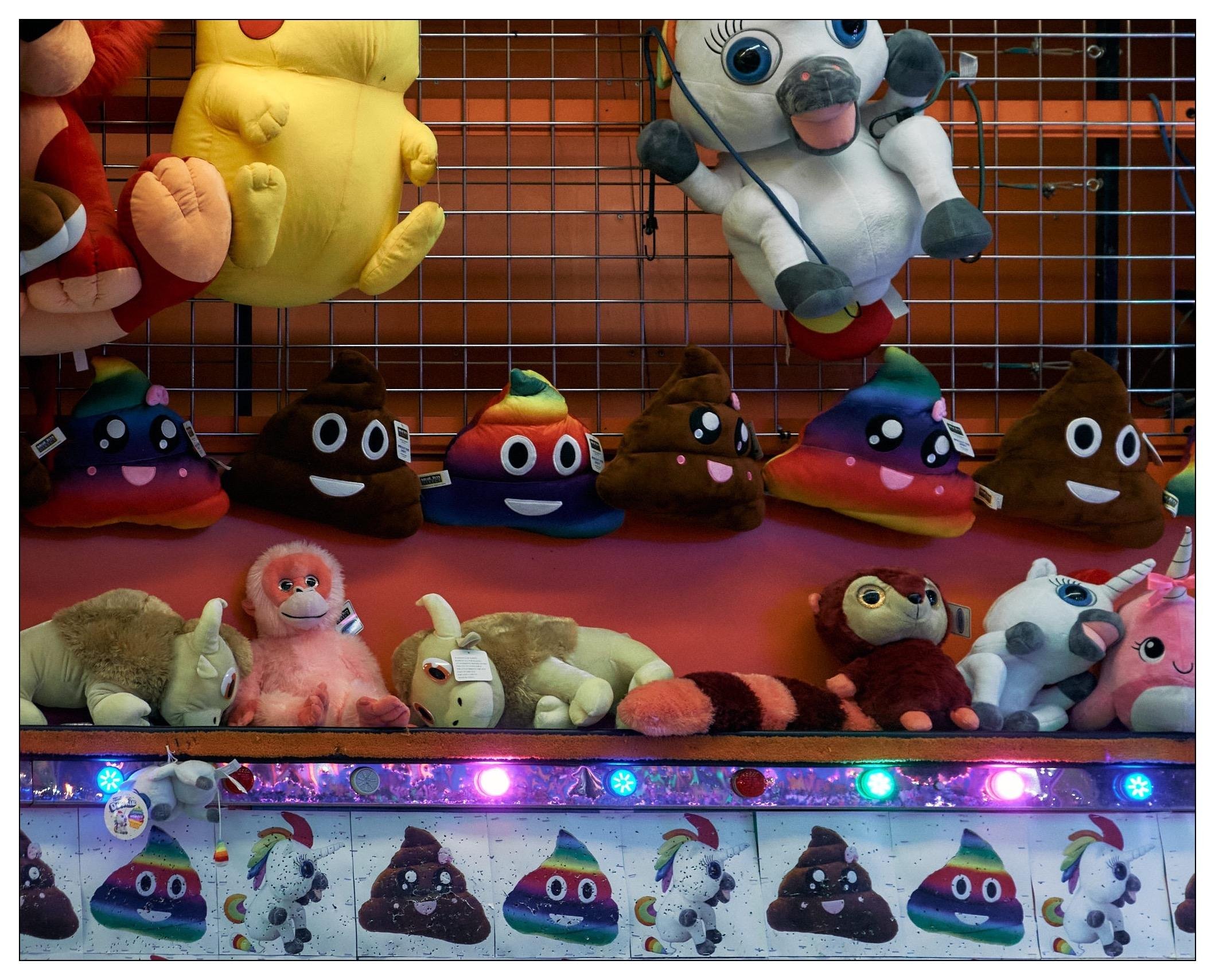 The image shows a variety of plush toys displayed in what appears to be a game or prize booth, possibly at a carnival or amusement park. There are several different types of toys:

1. On the upper left, there is a large plush resembling a creature with big eyes and large lips.
2. Hanging in the upper center, there's a plush toy that looks like a white horse or unicorn with wings, designed to look like it's parachuting.
3. Throughout the middle layer, there are numerous plush toys shaped like colorful poop emojis, each with different facial expressions. Some are smiling, while others are sticking out their tongues.
4. The bottom row includes several other animal plushies, such as a pink monkey, unicorns, an owl, and a striped toy resembling a caterpillar or a snake.

Overall, the toys are colorful and represent a mix of commonplace plush designs and unique, playful characters like the poop emojis. These items are typical to find as prizes at fairground games.