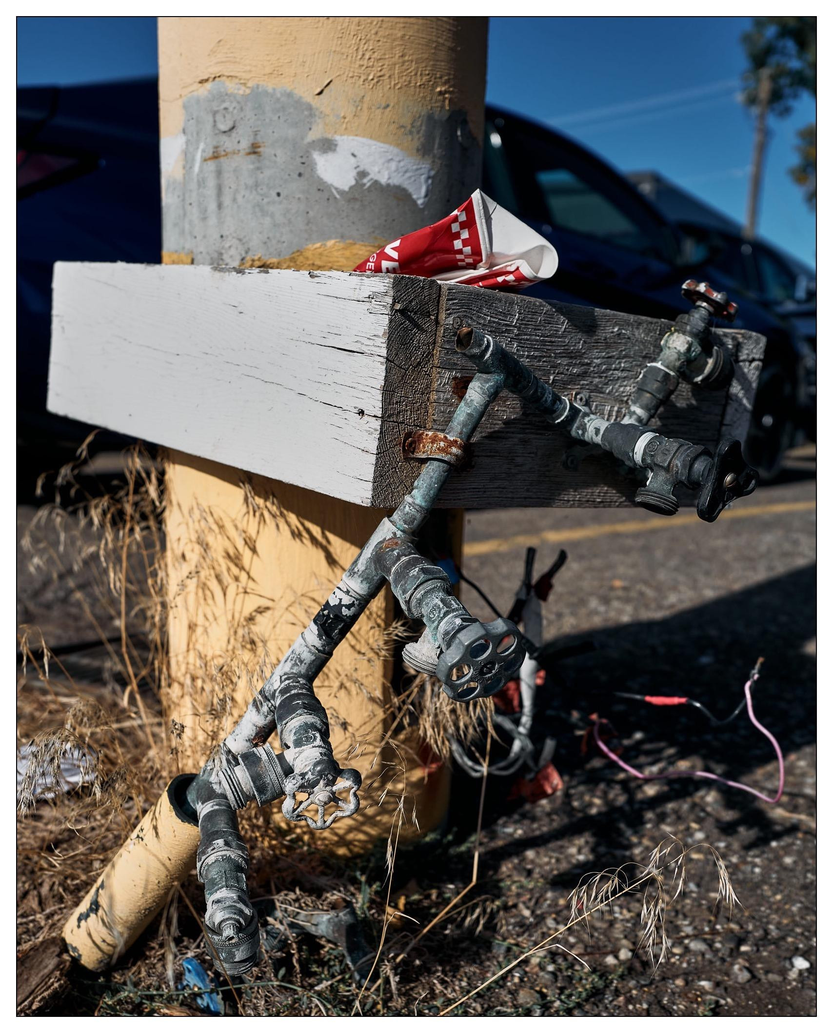 Vertical image of a weathered wooden and metal support structure with multiple elements. The structure features a white wooden beam strapped to a yellow cylindrical pole with worn paint and patches. Attached to the beam are several rusted metal pipes, valves, and faucets, arranged haphazardly. A discarded red and white striped paper cup is wedged into the beam. Dry grass and debris are visible at the base, with cars and clear blue sky in the background.