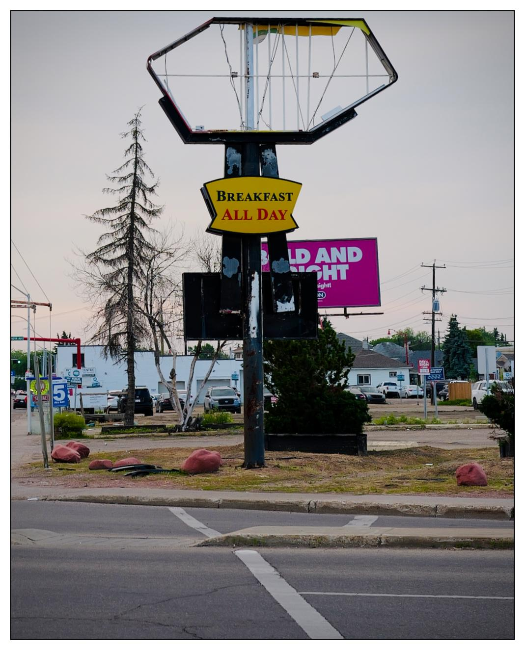 A broken, bent, hollow upper frame of a former Denny's sign, with a sub sign below fully intact that says "Breakfast all day".  There is no more Denny's here, it is an empty lot.
