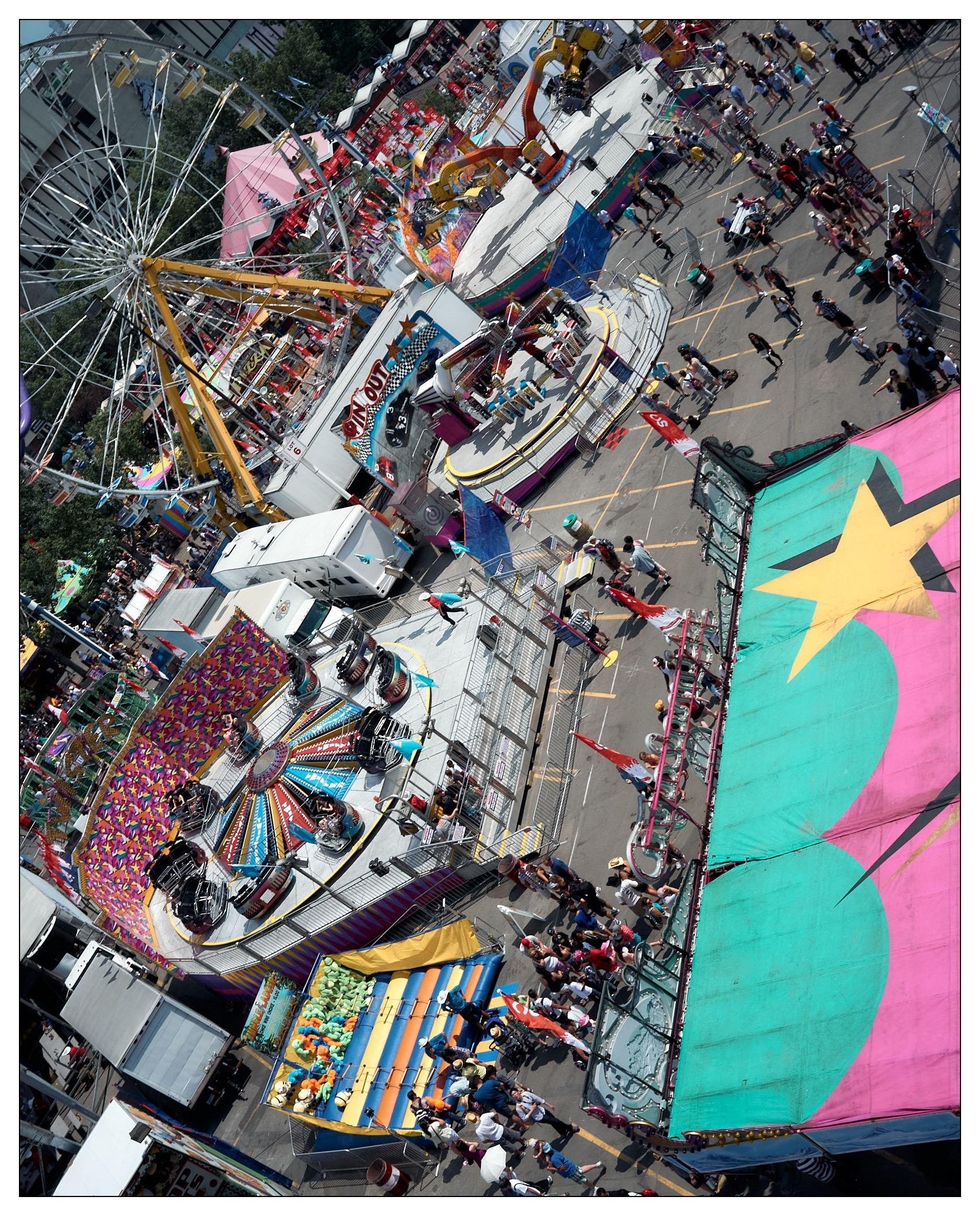 This image captures an aerial view of a bustling fair or amusement park. Prominently featured are various colorful amusement rides including a Ferris wheel, spinning chair swings, and other carnival attractions. Throngs of people can be seen walking around, and there are several game and food stalls line the walkways. The image gives a vibrant, festive feel, highlighted by the bright colors of the attractions and tents.