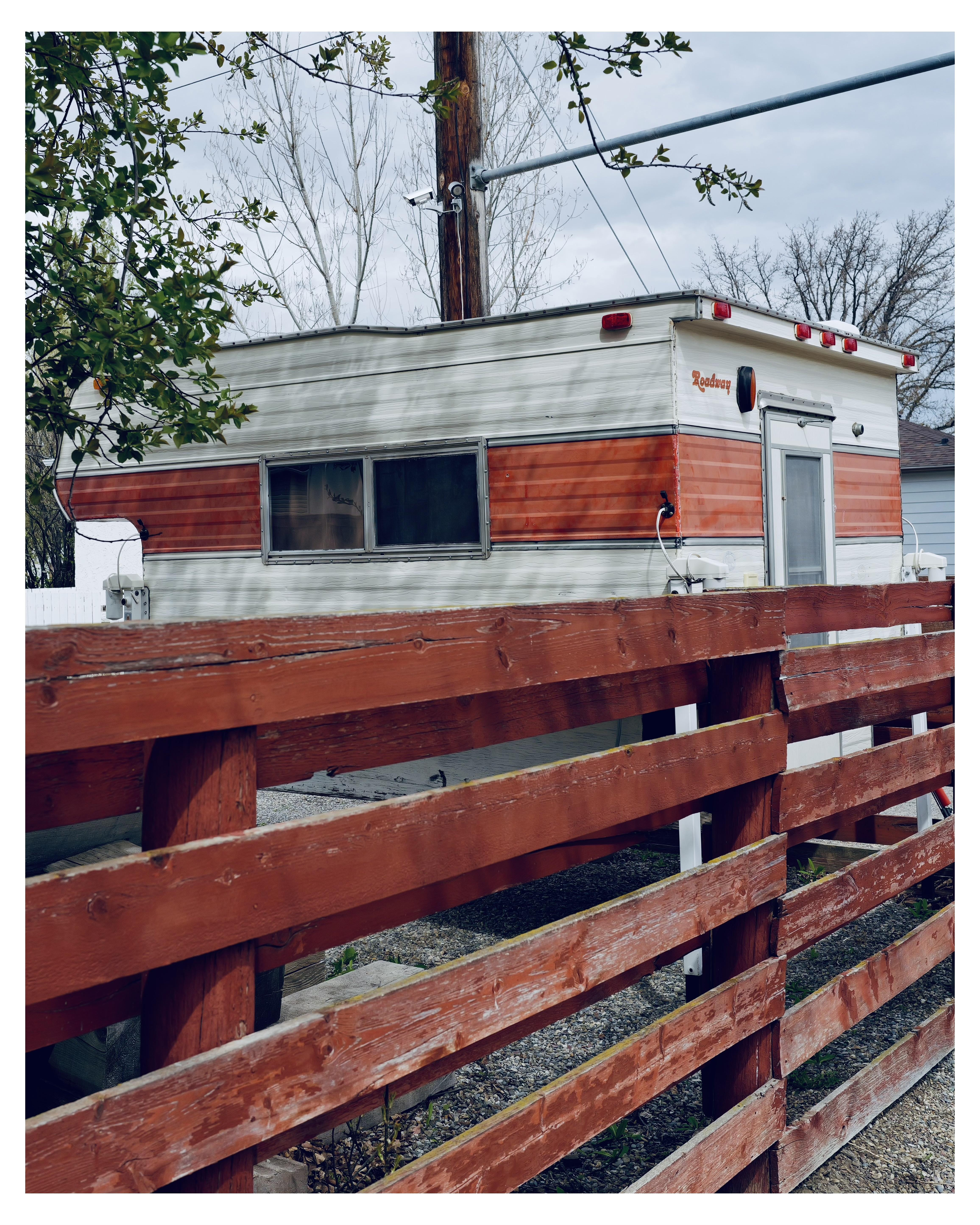 The image features an old, vintage-style truck camper with a two-toned body, primarily silver with a red stripe. The name "Roadway" is visible near the roof on the trailer side, possibly indicating the model or brand. This trailer is parked and elevated slightly above ground level, its surroundings partially hidden by a reddish wooden fence, which is weathered and shows signs of wear. The background includes leafy trees and a utility pole with wires, suggesting a residential or semi-rural setting. The overall atmosphere conveys a sense of quiet and stillness, typical of a peaceful neighborhood or a private property setup.