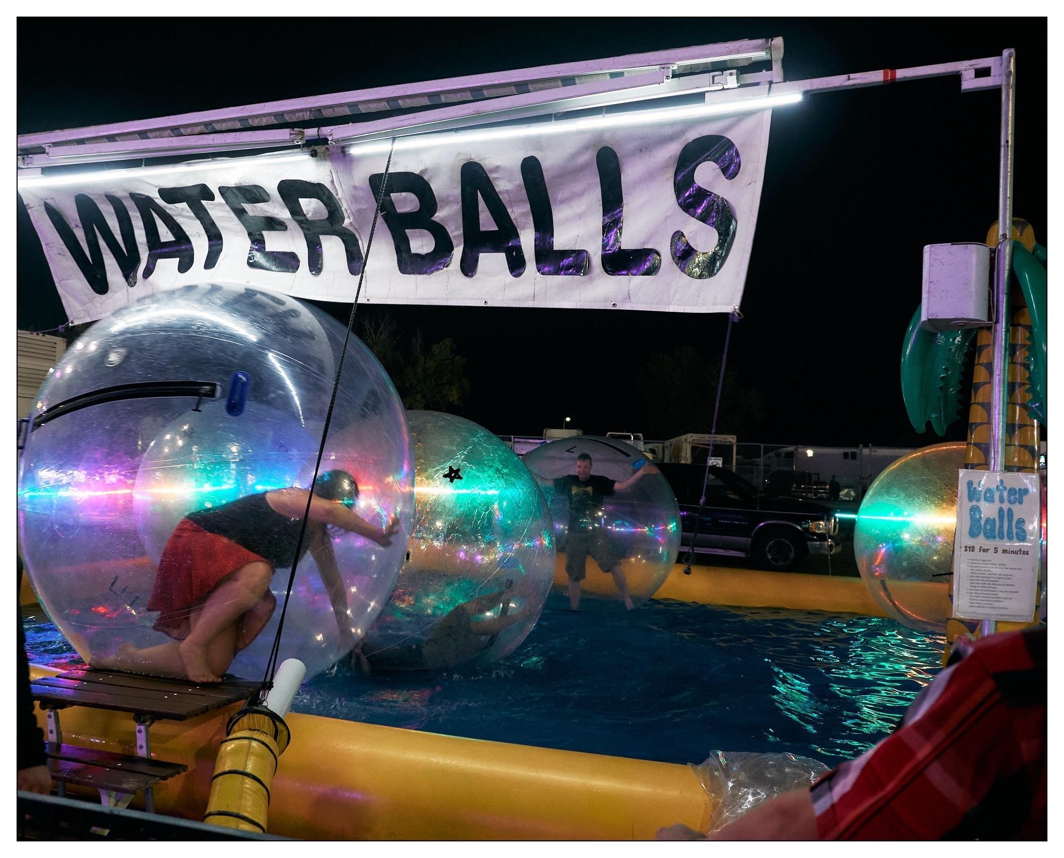 This image captures a fun and colorful scene at what appears to be a fair or amusement park. It shows an activity area titled "Water Balls," where people can go inside large inflatable balls that float on water. The image features two individuals inside these transparent orbs, floating in a small pool outlined with yellow barriers. One person is actively moving inside the ball, attempting to walk or roll it across the water, while the other appears to be standing still, possibly adjusting to the space or taking a moment to enjoy the activity. The area is illuminated by ambient and colorful lighting, which adds a lively and vibrant atmosphere to the scene.