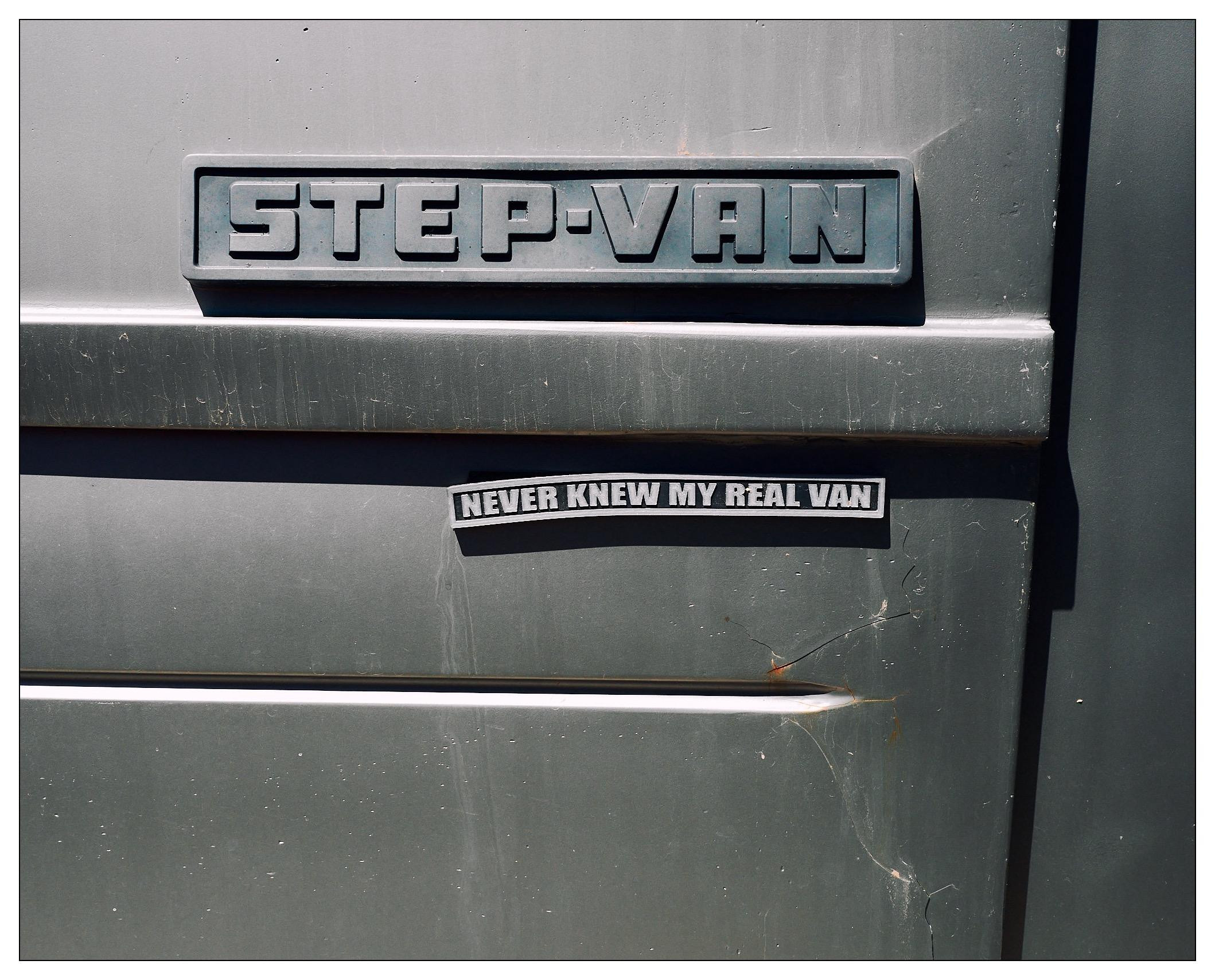 Close-up of a section of a gray van showing two emblems. The top emblem features the words "STEP-VAN" in block letters against a dark background. Below it, a smaller emblem reads "NEVER KNEW MY REAL VAN" against a gray background. Both emblems are set on a visible metallic surface with light scratches and dents.