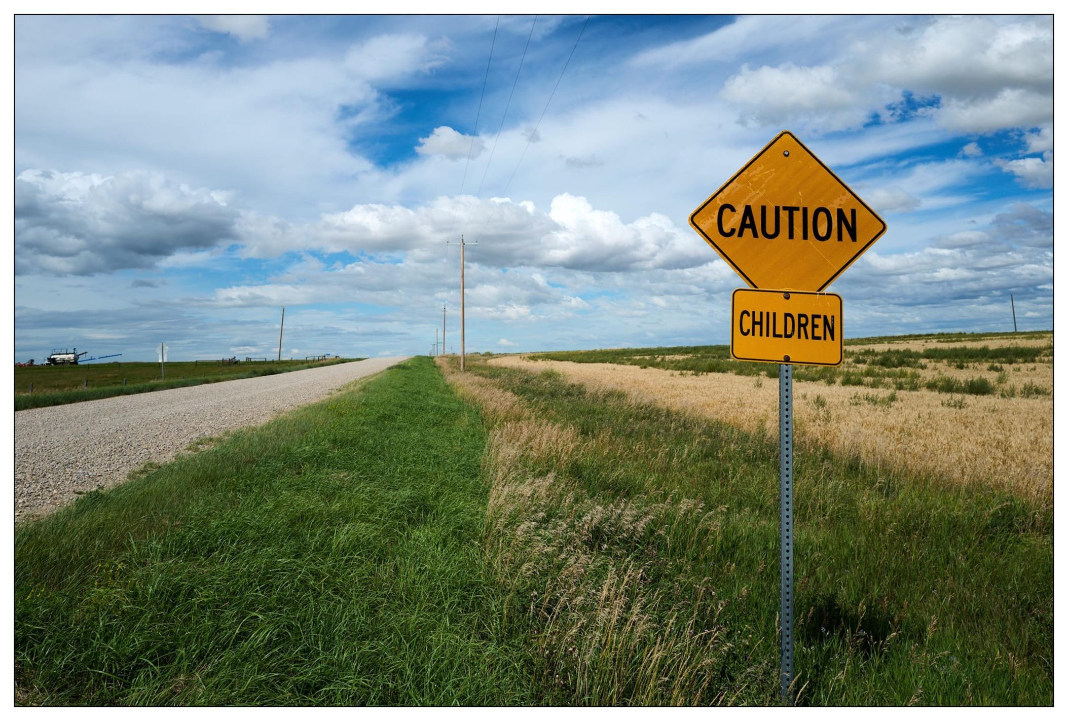 Yellow diamond-shaped road sign reading "CAUTION CHILDREN" on the right side of a rural road. The road stretches into the distance, flanked by green grass and golden fields under a cloudy sky. Utility poles line the left side of the road.