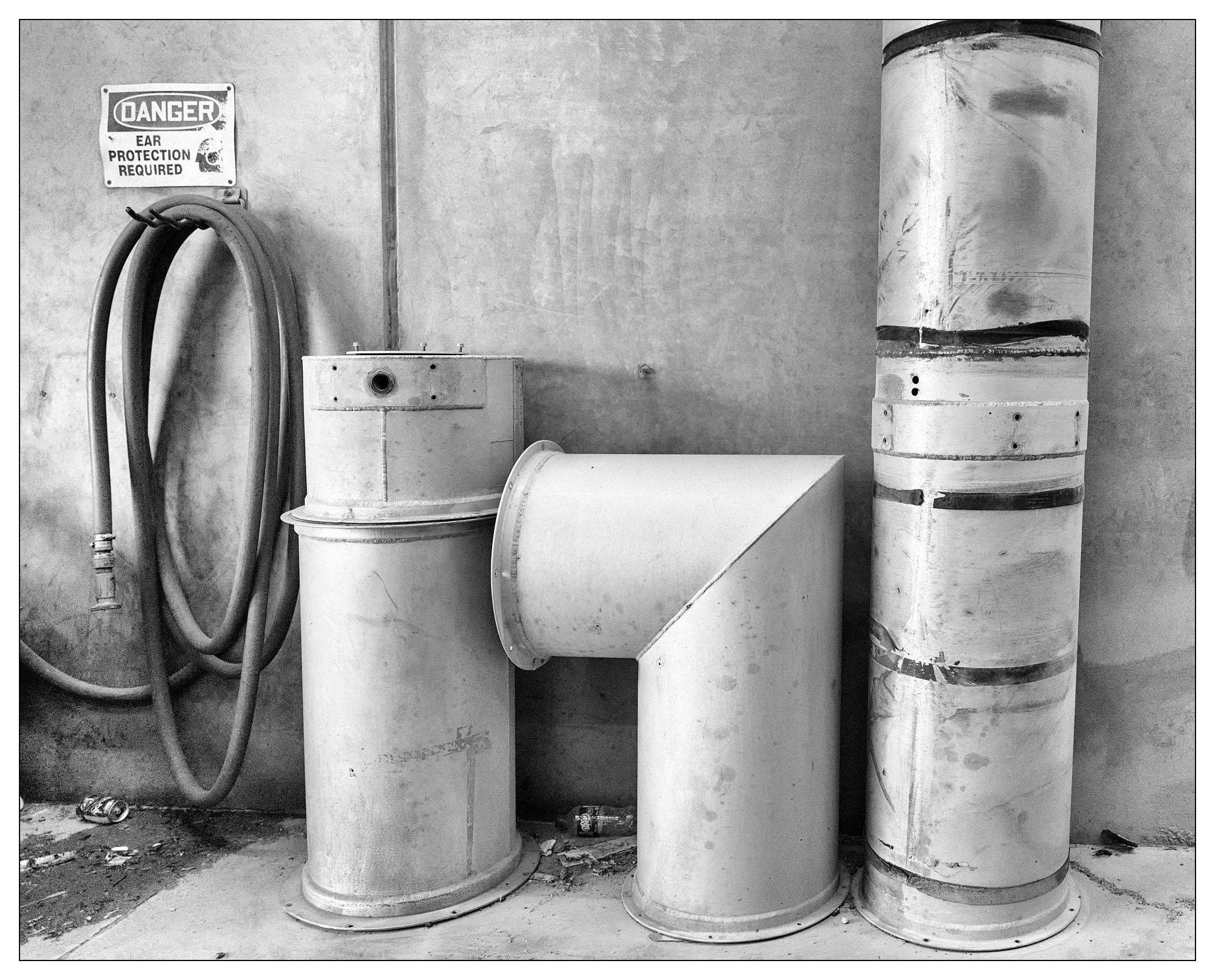Black and white photo featuring industrial metal ductwork and pipes against a concrete wall. A “Danger Ear Protection Required” sign is mounted on the wall above a coiled hose. The elements appear worn, with visible rust and markings.